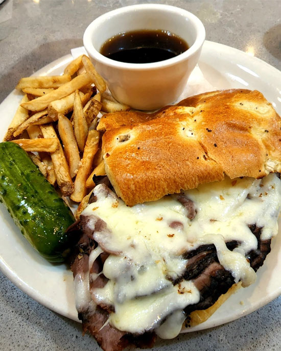 broadway deli dip. Roast beef topped with melted mozzarella on an onion roll served with a side of coleslaw and a pickle. Next to a cup on Au jus