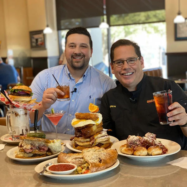 Photo of Max and Louie's restaurant owner Drew Glick smiling and sitting in front of various menu items with David Elder of Texas Eats television show