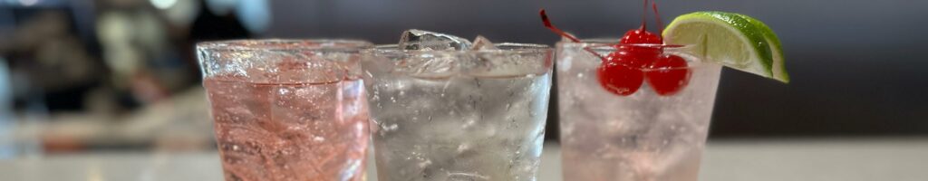closeup of three handmade fountain drinks. One is slightly pink with a garnish of three cherries and two limes, another with clear soda and the third with pink soda.