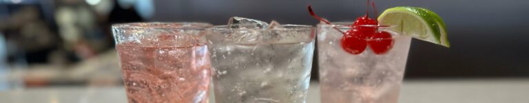 closeup of three handmade fountain drinks. One is slightly pink with a garnish of three cherries and two limes, another with clear soda and the third with pink soda.