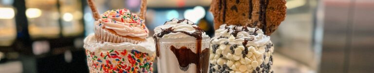 various milkshakes topped with sprinkles, a cupcake, pretzel rods, milk chocolate and white chocolate chips, chocolate sauce and a cookie