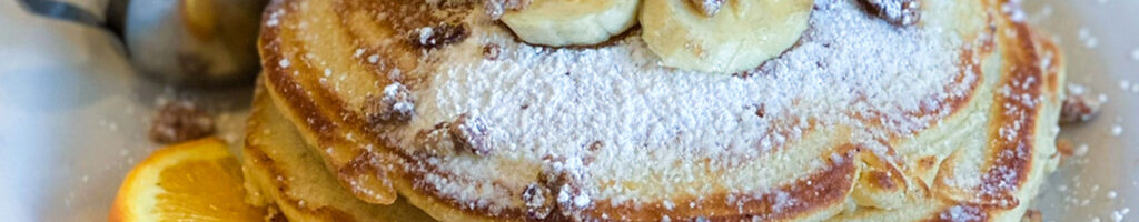 A stack of fluffy banana pancakes sprinkled with a light dusting of confectioner's sugar, generously drizzled with golden maple butter syrup, served at Max & Louie's NY Diner.